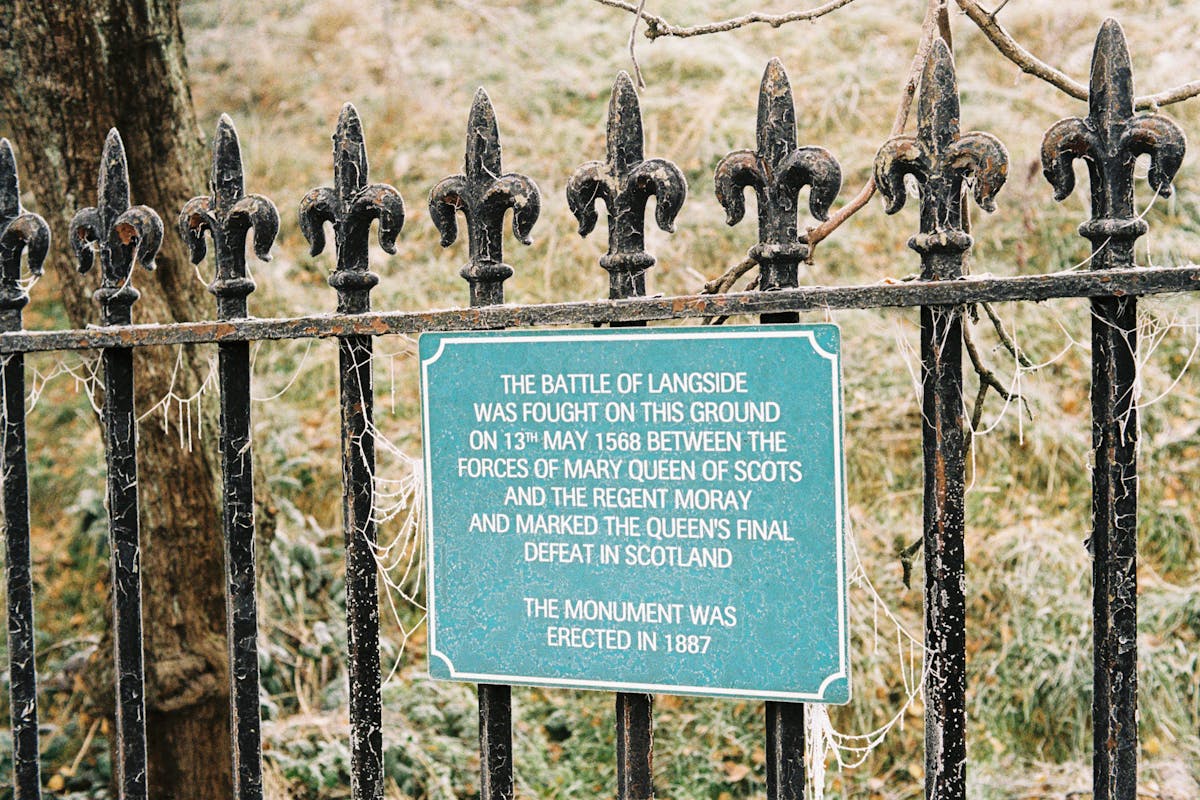 A sign on a fence, which reads 'The Battle of Langside was fought on this ground on 13th May 1568 between the forces of Mary Queen of Scots and the Regent Moray and marked the queen's final defeat in Scotland.'