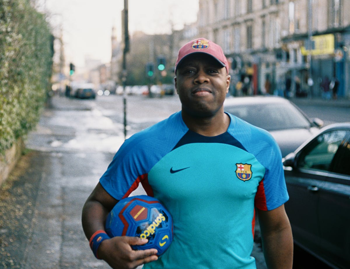 A man standing on the sidewalk in a soccer uniform, holding a soccer ball, looking at the camera