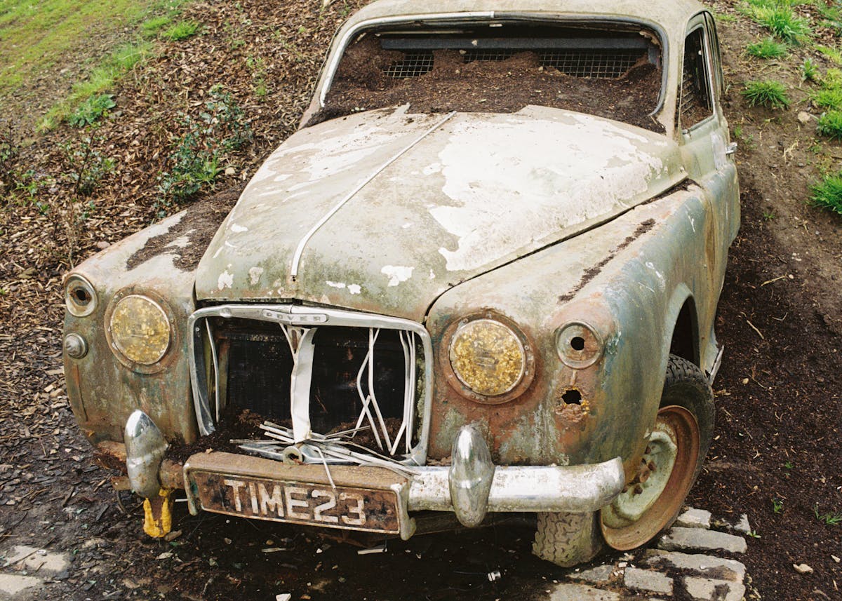 A junkyard car buried in diry with a license plate that says time.