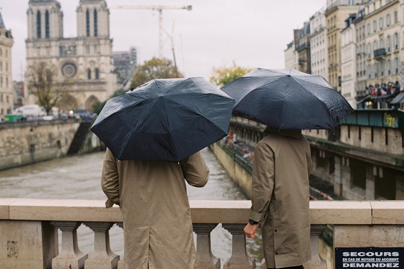 Two men in trench coats with umbrellas looking at Notre Dame cathedral.